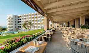 Ulkoterassi katettuin pöydin, viheralueineen ja taustalla monivärinen rakennus. (Tekoälyn luoma teksti)