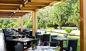 Ulkoravintola, jossa on katettuja pöytiä ja näkymä puutarhaan, jossa on palmuja. (Tekoälyn luoma teksti)