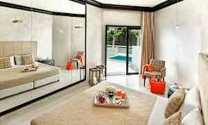 Hotellihuone, jossa suuri peiliseinä, beige vuodepeite ja tyynyt sekä parvekkeen ovi, joka johtaa uima-altaalle. (Tekoälyn luoma teksti)