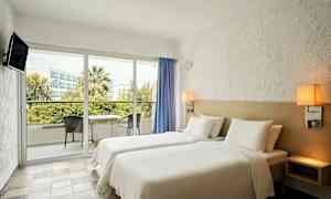 Hotellihuone, jossa on valkoiset vuodevaatteet ja kaksi erillistä vuodetta, parveke näkymällä puutarha-alueelle ja kaupungin taustalle. (Tekoälyn luoma teksti)