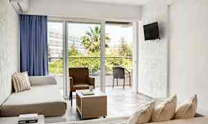 Valoisa hotelli huone, jossa on oleskelutila, sohvat, tuoli, sohvapöytä ja suuret ikkunat, jotka avautuvat parvekkeelle ulkokalusteilla vehreässä ympäristössä. (Tekoälyn luoma teksti)