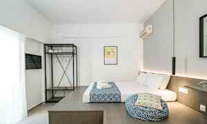 Moderni hotellihuone, jossa sänky, valkoiset ja siniset tekstiilit, avoin vaatekaappi, hyllyt ja vaateripustimet sekä seinään kiinnitetty ilmastointilaite. (Tekoälyn luoma teksti)