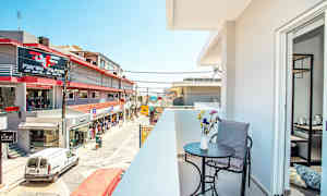 Parveke, jolla on pöytä, tuoli ja kukkia etualalla, sekä näkymä kadulle, jossa on liikkeitä ja rakennuksia. (Tekoälyn luoma teksti)