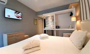 Kuvassa on hotellihuone, jossa on sänky, pyyhkeet, seinälle ripustettu televisio sekä varusteita kuten vaatekaappi, tallelokero ja vedenkeitin. (Tekoälyn luoma teksti)