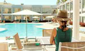 Henkilö istuu pöydän ääressä uima-altaan lähellä, jossa on aurinkovarjoja. Taustalla näkyy hotellirakennus. (Tekoälyn luoma teksti)