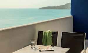 Kaksi tuolia, joiden yhteen on asetettu vihreä huopa, pöydän ääressä, jossa on vesilasi ja lehti, ja näkymä merelle. (Tekoälyn luoma teksti)