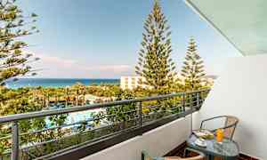 Parveke, jossa pöytä ja kaksi tuolia. Näkymä uima-altaalle ja vehreään alueeseen palmuineen sekä taustalla olevaan mereen. (Tekoälyn luoma teksti)