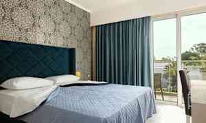 Hotellihuone, jossa on sänky, sininen peitto ja siniset verhot, sekä ikkuna, joka johtaa parvekkeelle. (Tekoälyn luoma teksti)