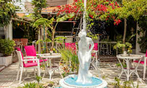 Suihkulähde, jossa on valkoinen naispatsas, vehreässä puutarhassa, jossa on pöytiä ja tuoleja. (Tekoälyn luoma teksti)