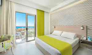 Hotellihuone, jossa sänky, vihreä päiväpeite, vaaleat huonekalut ja parveke merinäkymällä. (Tekoälyn luoma teksti)