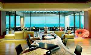 Moderni huone, jossa on suuret lasi-ikkunat ja näkymä merelle. Etualalla on pitkä puupöytä ja taustalla moderni istuinryhmä. (Tekoälyn luoma teksti)