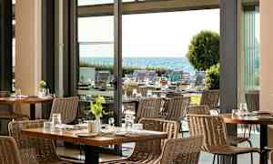 Näkymä moderniin ravintolaan, jossa punottuja tuoleja ja pöytiä, taustalla puutarha ja meri. (Tekoälyn luoma teksti)