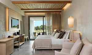 Moderni hotellihuone, jossa on parivuode, istuinryhmä ja kirjoituspöytä. Tekstiilit päästävät päivänvalon parvekkeen ovelta. (Tekoälyn luoma teksti)