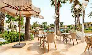 Ulkoiluterassi, jossa on pöytiä ja tuoleja, palmujen ja vehreyden ympäröimänä. Iso aurinkovarjo antaa varjoa. (Tekoälyn luoma teksti)