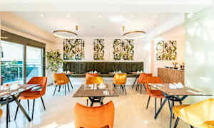 Ruokasali, jossa on oranssin ja keltaisen värisiä tuoleja sekä vihreä pehmustettu penkki. Pöydät on katettu ja koristeltu. Seinillä on kasviaiheisia kuvia. (Tekoälyn luoma teksti)