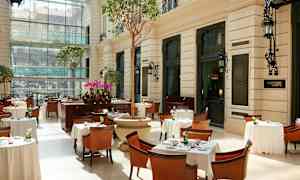 Valoisa ravintola, jossa on pöytiä, tuoleja ja kasveja lasitetussa ympäristössä. (Tekoälyn luoma teksti)