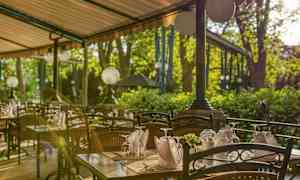 Ulkoravintola, jossa katettuja pöytiä ja tuoleja katoksen alla, taustalla vihreitä puita. (Tekoälyn luoma teksti)