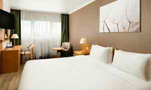 Hotellihuone, jossa on suuri parivuode, kirjoituspöytä, tuoli, nojatuoli ja ikkuna vaaleilla verhoilla. (Tekoälyn luoma teksti)