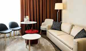 Olohuoneessa on vaalea sohva, pyöreä pöytä, kaksi tuolia ja ruskeat verhot. (Tekoälyn luoma teksti)