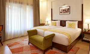 Hotellihuone, jossa on kaksi erillistä sänkyä, vihreä penkki ja vaalea ikkunaverho. (Tekoälyn luoma teksti)