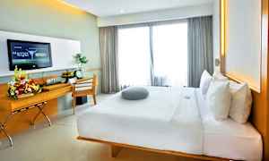 Moderni hotellihuone, jossa suuri mukava sänky, kirjoituspöytä kukka-asetelmalla ja seinällä televisio. Ikkunoista tulee luonnonvaloa läpi vaaleiden verhojen. (Tekoälyn luoma teksti)