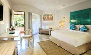 Valoisa hotellihuone, jossa on iso sänky, sohva ja työpöytä. Huoneessa on myös parveke, josta on näkymä vehreyteen. (Tekoälyn luoma teksti)