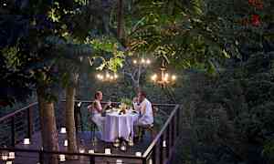 Romanttinen illallinen katetaan puuterassille vehreän kasvillisuuden ympäröimänä, valaistuna kynttilöillä ja kattokruunuilla. (Tekoälyn luoma teksti)