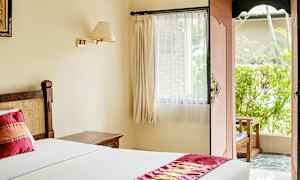 Valoisa hotellihuone, jossa on sänky valkoisilla vuodevaatteilla ja koristeellinen punainen kaitaliina. Ikkuna verhoineen ja avoin ovi johtavat ulkotilaan, jossa on vehreyttä. (Tekoälyn luoma teksti)