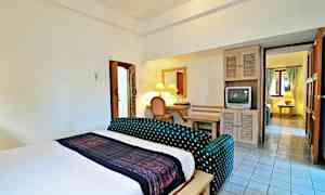 Hotellihuone, jossa on suuri sänky, pieni istuinryhmä, työpöytä peilillä ja vanha televisio. Kylpyhuone on vasemmalla, ja taustalla näkyy yhteys toiseen huoneeseen. (Tekoälyn luoma teksti)