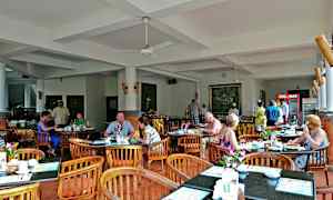 Ravintolatila, jossa on pöytiä ja tuoleja, ja ihmiset syövät. Sisustus on vaalea valkoisine seinineen ja kattoineen. (Tekoälyn luoma teksti)