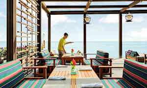 Ravintola meren äärellä puupöydillä ja raitatyynyin varustetuilla tuoleilla. (Tekoälyn luoma teksti)