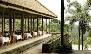 Ulkona sijaitseva ravintola, jossa on olkikatto ja katettuja pöytiä, rehevän vihreyden ympäröimänä. (Tekoälyn luoma teksti)