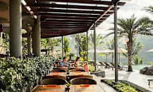 Ulkoterassi, jossa on puupöytiä ja -tuoleja, puulaudoista tehty katto ja vihreitä kasveja. Taustalla näkyy uima-allas ja palmuja. (Tekoälyn luoma teksti)