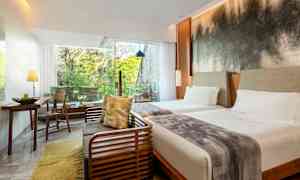 Hotellihuone, jossa on kaksi vuodetta, oleskelutila ja suuret ikkunat, jotka päästävät valoa sisään ja tarjoavat näkymän vehreisiin puihin. (Tekoälyn luoma teksti)