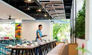 Ravintolan tila, jossa on pöytiä ja tuoleja, ja henkilö kantaa tarjotinta kuppien kanssa. (Tekoälyn luoma teksti)