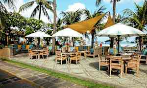 Ulkoilmaravintola meren rannalla, jossa on puupöytiä, tuoleja, varjokatoksia ja palmuja. (Tekoälyn luoma teksti)