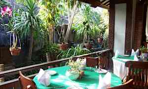 Ulkoalue, jossa on pöytiä ja tuoleja terassilla. Pöytiä on koristeltu vihreillä liinoilla, lautasilla ja lautasliinoilla. Taustalla näkyy vihreää kasvillisuutta ja roikkuvia kasveja. (Tekoälyn luoma teksti)