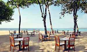 Puupöytiä ja -tuoleja ulkona hiekkarannalla, taustalla meri ja puita. (Tekoälyn luoma teksti)