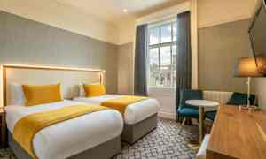 Kaksi vuodetta, joissa keltaiset päiväpeitot ja tyynyt, huoneessa on pöytä, tuolit ja ikkuna. (Tekoälyn luoma teksti)