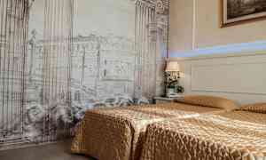 Hotellihuone, jossa on kaksi sänkyä kultaisen väristen päiväpeitteiden peitossa. Taustaseinässä on arkkitehtonisia pylväitä ja rakennuksia esittävä teos. Yöpöydällä on yölamppu ja kasvi. (Tekoälyn luoma teksti)