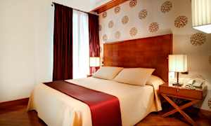 Makuuhuone, jossa on suuri beige- ja viininpunaisilla vuodevaatteilla peitetty sänky, kaksi yöpöytää valaisimilla ja koristeellinen tapetti seinällä. (Tekoälyn luoma teksti)