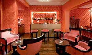 Kuva, joka esittää eleganttia aluetta sisustettuna cocktailbaariksi tummilla huonekaluilla, vaaleanpunaisilla tyynyillä ja hyllyllä olevilla pulloilla. (Tekoälyn luoma teksti)