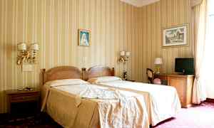 Hotellihuone, jossa on kaksi yhden hengen sänkyä beigeillä vuodevaatteilla, koristeltuja seiniä tauluilla ja huonekaluja, kuten pöytiä, tuoleja ja televisio. (Tekoälyn luoma teksti)