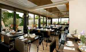 Valoisa ravintola, jossa on katetut pöydät, koristeltuna kukilla ja ikkunat, jotka paljastavat ulkona olevan ympäristön. (Tekoälyn luoma teksti)