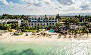 Ranta, jossa kirkasta vettä, aurinkovarjoja ja aurinkotuoleja, ympärillä hotelli ja vihreää. (Tekoälyn luoma teksti)