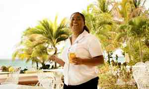 Henkilö pitelee kahta lasia, toista viinin ja toista mehun kanssa, ulkona pöydän äärellä lähellä rantaa ja palmujen ympäröimänä. (Tekoälyn luoma teksti)