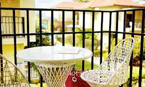 Parveke, jossa on kaksi valkoista punottua tuolia ja pyöreä pöytä. Pöydällä on kirja. Näkymä viheralueille ja rakennuksiin taustalla. (Tekoälyn luoma teksti)