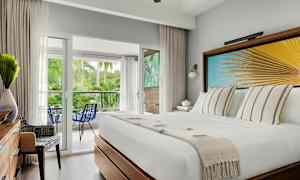 Hotellihuone, jossa valkoinen vuode, koristeellinen seinätaideteos palmulehdillä, näkymä parvekkeelle vihreässä ympäristössä. (Tekoälyn luoma teksti)