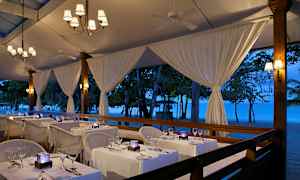 Ravintola, jossa on pöytiä valkoisilla liinoilla rannalla iltavalaistun teltan alla. (Tekoälyn luoma teksti)