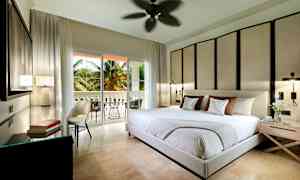 Moderni makuuhuone, jossa iso sänky, koristeellinen seinäpaneeli ja käynti parvekkeelle, ulkona palmuja. (Tekoälyn luoma teksti)